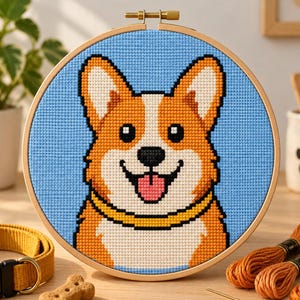 Puede incluir: Un bastidor de bordado circular de madera que contiene un patrón de punto de cruz de un perro Corgi. El perro tiene pelaje naranja y blanco, nariz negra y lengua rosa, sobre un fondo azul claro. El Corgi lleva un collar amarillo sólido.
