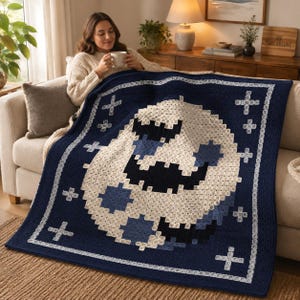 Op de afbeelding: Een donkerblauwe gehaakte deken met een groot, centraal gepixeld maanontwerp in tinten crème, lichtblauw en donkerblauw. De deken is omrand met een wit geometrisch patroon en bevat kleine witte kruisvormige accenten.