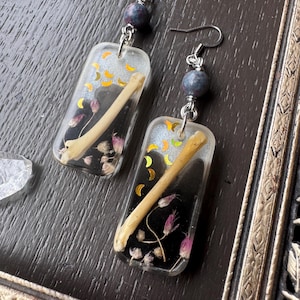 May include: A pair of rectangular resin earrings featuring a black and clear background with embedded small bones, dried purple flowers, and gold crescent moon decals. Each earring hangs from a silver-toned hook with a single blue-grey bead.
