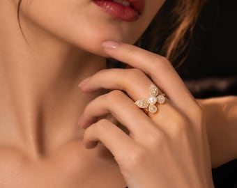 May include: A gold ring featuring a butterfly-shaped design adorned with small clear stones and a single central white pearl. The ring is worn on a finger, showcasing the detailed metalwork and the arrangement of the stones and pearl.