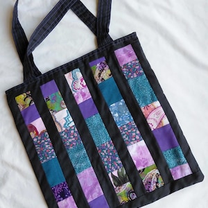 May include: A rectangular tote bag featuring a patchwork design of vertical fabric strips in shades of purple, teal, and blue, interspersed with solid black fabric columns. The bag includes two long black shoulder straps with subtle stitching.