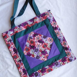 May include: A tote bag featuring a central diamond-shaped floral fabric panel in shades of red, purple, and white. This panel is framed by solid purple fabric and a dark teal border, with an outer floral print border and matching dark teal straps.