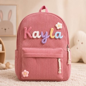 Puede incluir: Una mochila de pana rosa con el nombre Kayla en letras multicolores, texturizadas y similares a hilo en la parte delantera. La bolsa incluye un bolsillo frontal con cremallera, tiradores de borlas blancas y dos pequeñas flores de ganchillo.
