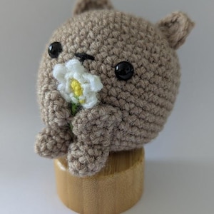 May include: A round, light brown crocheted animal figure with small ears, black plastic eyes, and a stitched nose. The figure holds a small white crocheted flower with a yellow centre in its front paws and sits atop a cylindrical wooden base.