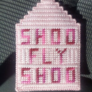 May include: A handmade, pink needlepoint ornament with the words "SHO FLY SHO" in a darker pink. The ornament is shaped like a house with a stepped roof and a metal loop for hanging.