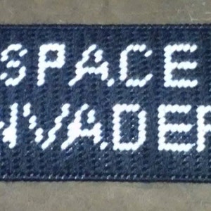 May include: A black and white crocheted doormat with the words "SPACE INVADER" in white.