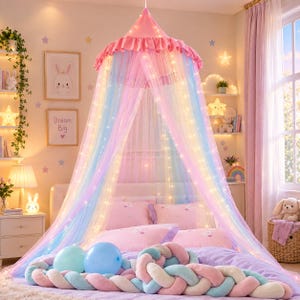May include: A pastel rainbow bed canopy made of sheer tulle fabric with a ruffled top, illuminated by integrated string lights. The canopy hangs over a bed featuring pink bedding and a braided, multi-colored plush knot pillow.