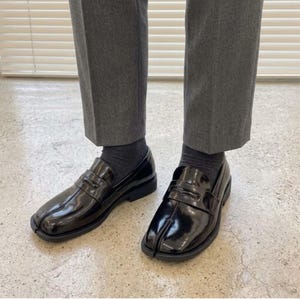 May include: A pair of black patent leather loafers featuring a distinctive split-toe design, a classic penny strap across the vamp, and a low block heel. The shoes are worn with dark gray socks and gray trousers, showcasing a polished finish.