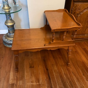Puede incluir: Una mesa auxiliar de madera de dos niveles con un acabado marrón medio. El nivel inferior es una superficie rectangular, mientras que el nivel superior elevado está sostenido por cuatro husillos torneados y detalles festoneados.