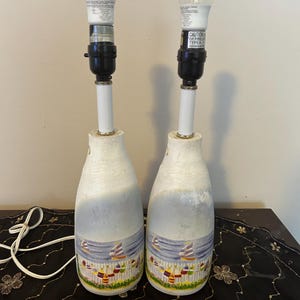 May include: A pair of table lamps featuring bottle-shaped bases with a painted nautical scene of sailboats on water and colourful buoys hanging on a white picket fence. Each lamp has a black socket, a white stem, and a clear light bulb on top.