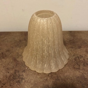 May include: A bell-shaped glass light shade featuring a translucent beige hue with a textured, mottled white finish. The shade has a fluted, scalloped bottom edge and a circular opening at the top for mounting to a light fixture.