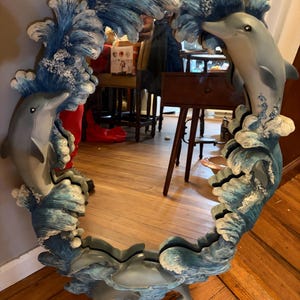 May include: An oval mirror featuring a decorative frame sculpted with three-dimensional dolphins leaping through crashing blue and white ocean waves. The frame is painted in shades of blue, white, and grey, creating a dynamic aquatic scene.