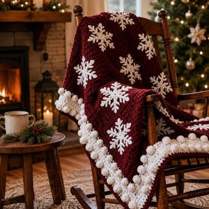 May include: A deep burgundy crochet blanket featuring white snowflake motifs and a white pom-pom border draped over a wooden rocking chair. The blanket has a thick, textured yarn construction with repeating snowflake patterns across the fabric.