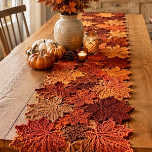May include: A long table runner crafted from interconnected crochet maple leaves in shades of orange, red, brown, and yellow. The runner features a detailed leaf pattern and is displayed on a wooden table with a ceramic vase and small pumpkins.