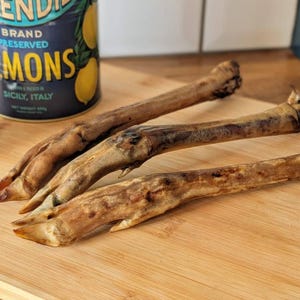 May include: Three dried, light brown animal trotters with visible hooves and textured skin, arranged diagonally. In the background, a blue cylindrical tin labelled SPLENDID BRAND PRESERVED LEMONS, GROWN & PACKED IN SICILY, ITALY, NET WEIGHT 500g.