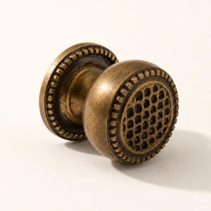 May include: A round cabinet knob with an antique brass finish, featuring a textured honeycomb pattern on the front face and a decorative beaded border around the edge of the circular base and the central knob component.