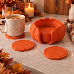 Pode incluir: Um conjunto de bases para copos redondas cor de laranja com um suporte a condizer em forma de abóbora. Uma base está sob uma caneca bege salpicada com uma bebida quente, enquanto duas bases adicionais estão planas ao lado do suporte.