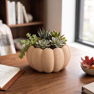 May include: A light beige, pumpkin-shaped ceramic planter filled with a variety of green succulents, including a trailing sedum and a striped haworthia, sitting on a wooden surface next to an open notebook and a small bowl of autumn leaves.