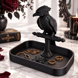 May include: A matte black jewelry tray featuring a central perch with a sculpted raven figure. The tray holds a gold-toned chain necklace with a dark stone pendant, a small black decorative emblem, and two gold-toned metal rings.