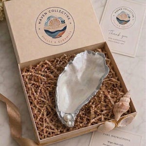 May include: A gift box containing a silver-painted oyster shell dish adorned with two white pearls. The box includes brown paper filler, a Haven Collective logo, a thank you card, and a care instructions card. A gold ribbon is tied around the box.
