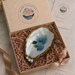 May include: A decorative oyster shell dish with a white interior, blue floral decal, and gold-painted edges, accented with three small white pearls. It is nestled in a cardboard gift box filled with shredded paper, accompanied by a branded card.
