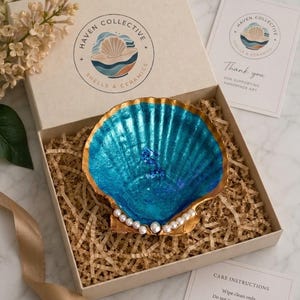 May include: A decorative seashell dish with a vibrant metallic blue interior and gold-painted edges, adorned with a row of small white faux pearls at the base. The shell is nestled in a gift box filled with shredded paper, accompanied by a brand card.