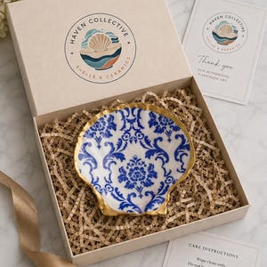 May include: A decorative shell dish with a blue and white damask pattern and a gold-painted edge, nestled in a beige gift box with paper filler. The box lid features a Haven Collective logo, and includes a thank you card and care instructions.
