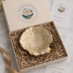 May include: A white seashell dish featuring a gold mandala pattern, nestled in a beige gift box with brown paper filler. The box lid displays the Haven Collective logo. Included are a thank you card and a care instructions card.