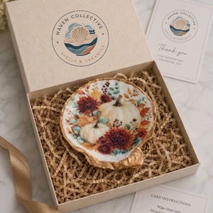 May include: A decorative seashell dish with a painted autumn scene featuring white pumpkins and dark orange flowers, finished with a gold-painted edge. The dish is nestled in a beige gift box with shredded paper filler, a logo, and cards.