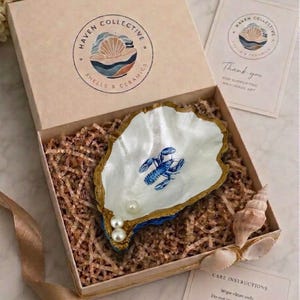 May include: A decorative oyster shell dish with a painted blue lobster and three white pearls inside, nestled in a kraft paper gift box filled with shredded paper filler. The box lid features a Haven Collective logo and includes a thank you card.