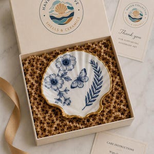 May include: A white shell-shaped ceramic dish with a gold-painted rim, featuring blue floral and butterfly illustrations. It is presented in a branded box with crinkle paper, a thank you card, and a care instruction card from Haven Collective.