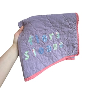Peut inclure: Une couverture matelassée lavande avec une bordure rose corail et des lettres en tissu appliquées formant le nom Flora Sloane. Les lettres sont confectionnées dans divers tissus à motifs, notamment des rayures et des carreaux bleus, verts et rouges.