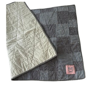 May include: A patchwork quilt featuring alternating squares of blue and white striped fabric and small black and white gingham check fabric. The reverse side is a solid off-white fabric with visible stitching. A pink label reads A&E.