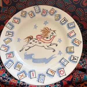 May include: A white ceramic plate featuring a central illustration of a galloping spotted horse with a flowing mane and a red scarf. The rim is decorated with a border of individual letters of the alphabet enclosed in black-outlined squares.