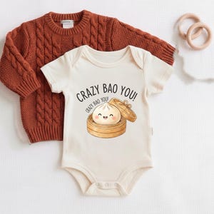 May include: A cream-coloured short-sleeve bodysuit featuring a graphic of a smiling bao bun in a bamboo steamer with the text "CRAZY BAO YOU!" layered over a rust-coloured long-sleeve cable knit jumper and two light wooden teething rings.