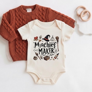 May include: A cream-colored short-sleeve bodysuit featuring the text "MISCHIEF MAKER" in black, surrounded by illustrations of a witch hat, broom, potion bottle, chest, and glasses. A rust-colored cable-knit sweater is layered behind the bodysuit.