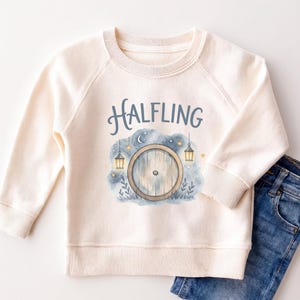 May include: A cream-colored long-sleeve sweatshirt featuring a graphic print of a round wooden door, two hanging lanterns, stars, and a crescent moon. The text "HALFLING" is printed in blue above the illustration, which is set against a blue watercolor background.