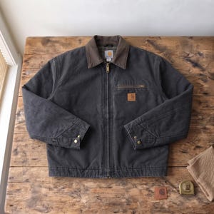 May include: A charcoal grey Carhartt jacket with a brown corduroy collar, a full-length front zip, and a zipped chest pocket. The jacket features a square logo patch on the chest and adjustable snap-button cuffs. A measuring tape is visible.