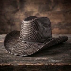 May include: A dark brown cowboy hat featuring a textured snakeskin pattern across the entire surface. The hat includes a matching narrow band wrapped around the base of the crown and a wide, curved brim that slopes downward on the sides.