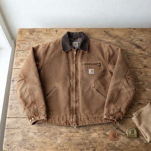 May include: A brown Carhartt jacket with a dark brown corduroy collar, a front zip fastening, and a chest pocket with a zip. The jacket features a square logo patch on the chest, two lower slanted pockets, and buttoned cuffs on the sleeves.
