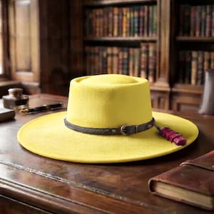 Puede incluir: Un sombrero amarillo brillante de ala plana y copa plana. El sombrero presenta una banda de cuero trenzado de color marrón oscuro con una hebilla de metal y un pequeño detalle de pluma de color rojo oscuro en el lateral de la banda.