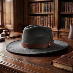 Può includere: Un cappello fedora in feltro grigio antracite con tesa larga e una fascia in pelle marrone liscia attorno alla corona. La fascia in pelle è fissata con un piccolo dettaglio a borchia metallica sul lato, conferendo al cappello un aspetto pulito.
