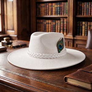 May include: A white wide-brimmed fedora hat featuring a braided white hatband and a single peacock feather accent tucked into the side. The hat has a structured crown with a central crease and a flat, circular brim, presented in a neutral tone.