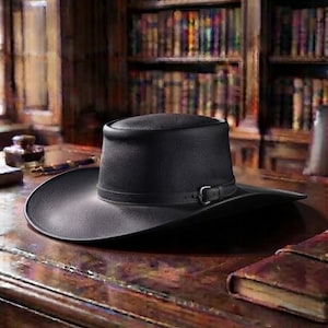 May include: A black leather hat with a flat top and a wide, curved brim. The hat features a thin leather band around the base of the crown, secured with a small, dark metal buckle on the side. The texture of the leather appears grained and matte.