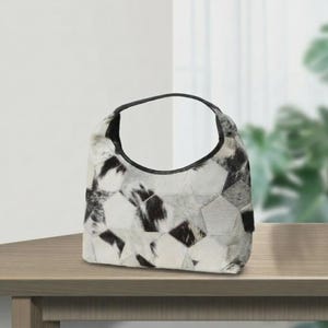 May include: A hobo-style handbag featuring a patchwork design of cowhide panels in black, white, and gray. The bag has a curved top opening with a solid black leather trim along the edge and a single integrated handle for carrying.