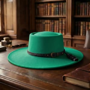 Puede incluir: Un sombrero de fieltro verde vibrante con copa plana y ala ancha. El sombrero presenta una banda de cuero marrón oscuro alrededor de la base, acentuada con una pequeña hebilla de metal y una borla de cuero decorativa en el lateral.