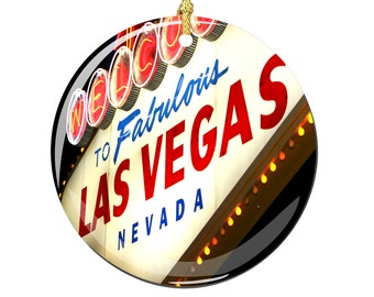 May include: Round ceramic ornament featuring a vintage-style neon sign that reads "Welcome to Fabulous Las Vegas, Nevada". The sign is white with red and blue lettering and has a black background.