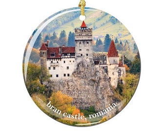 May include: A round glass ornament featuring a photo of Bran Castle in Romania. The castle is a white stone structure with red tile roofs, set against a backdrop of green trees and hills. The text "bran castle, romania" is printed in white at the bottom of the ornament.