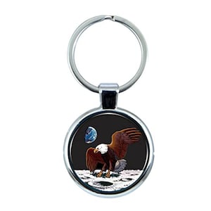 Puede incluir: Un llavero de plata con un dije redondo de plata. El dije presenta un fondo negro con un águila calva de pie en la luna. El águila mira a la Tierra, que es visible en el fondo.