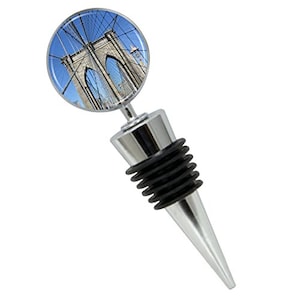 May include: A silver bottle stopper with a round, clear glass top featuring a photo of the Brooklyn Bridge. The stopper has a black rubber seal and a pointed tip.